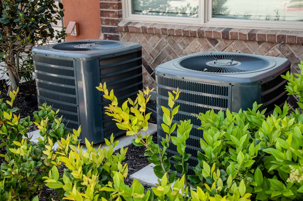Two outdoor HVAC units sitting on concrete slabs over mulch and behind green bushes under a window